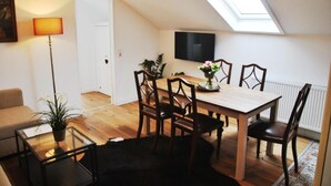 A cozy living room with a wooden dining table, chairs, a sofa, a glass coffee table with a plant, a floor lamp, and a TV mounted on the wall. - Gutshaus beim Zeilinger Schlössl (Knittelfeld)