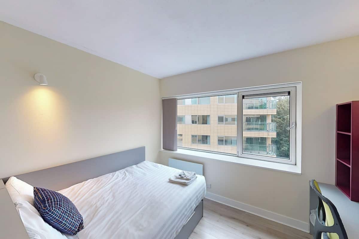 Un dormitorio con cama, ventana y vistas a edificios.