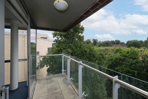 Un balcon doté d’une balustrade en verre, d’un sol carrelé et offrant une vue sur les arbres et les bâtiments en arrière-plan.