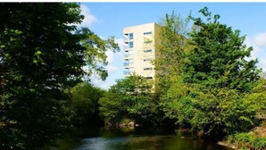 A modern building with a river in the foreground and lush greenery around. - Victoria Mills (Cork)