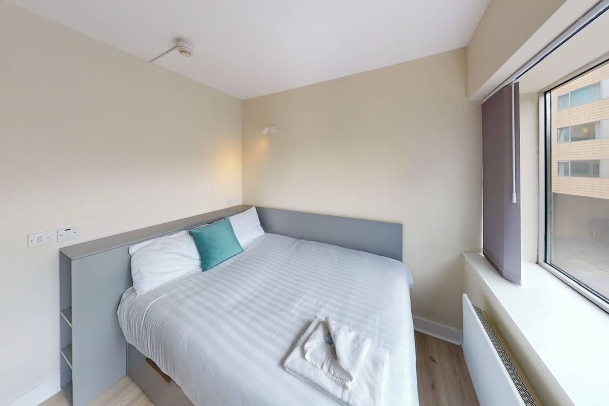 Una cama bien hecha con un cabecero gris, almohada blanca y una almohada de acento color turquesa. Se ve una ventana con vista a un edificio.