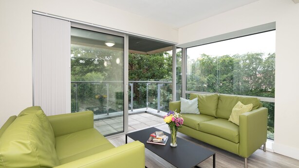 A modern living room with a green sofa, a black coffee table, and a glass door leading to a balcony with a view of trees. - Victoria Mills (Cork)