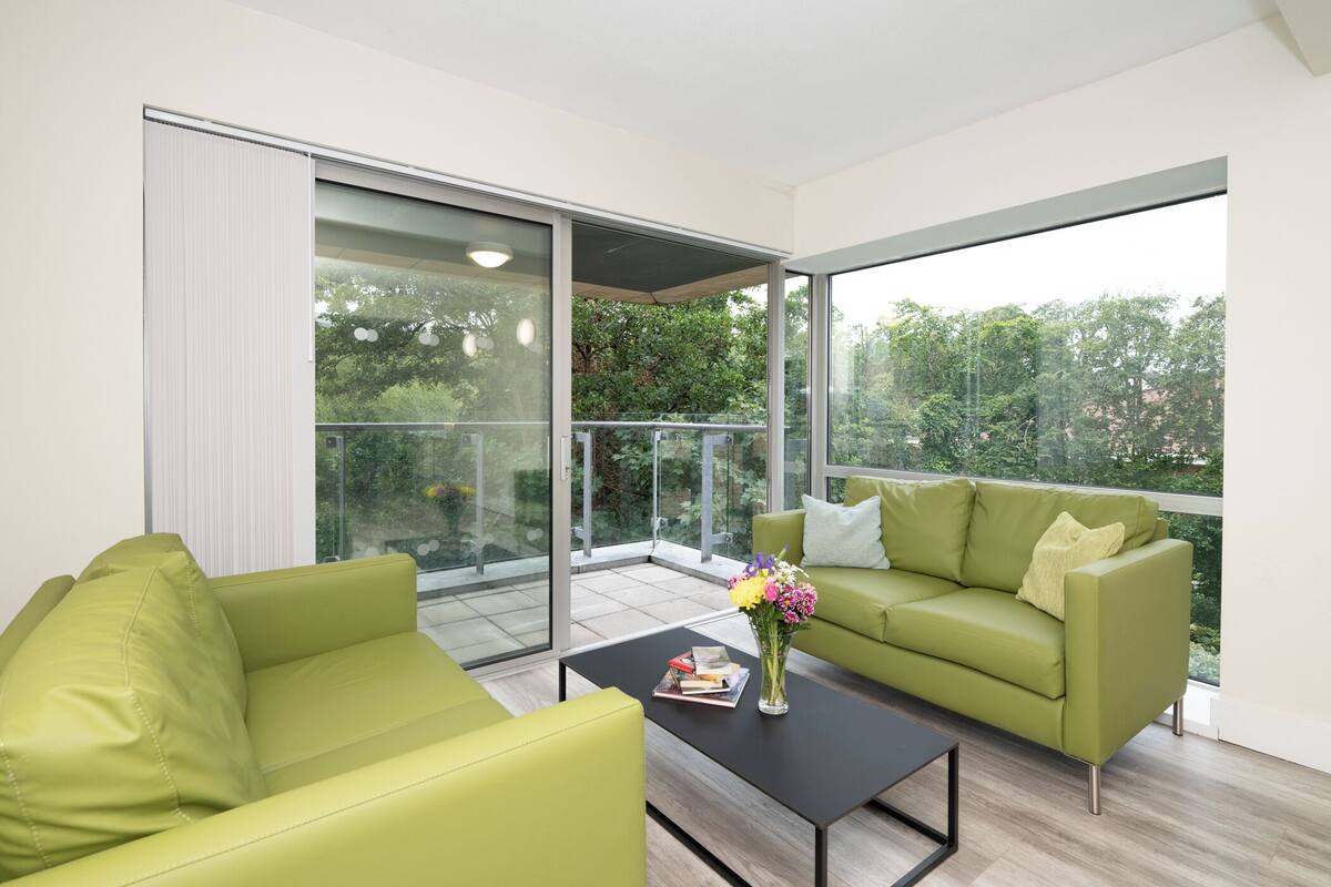 Un salón moderno con un sofá verde, una mesa de centro negra y un ventanal de vidrio que da a un balcón con vistas a árboles.