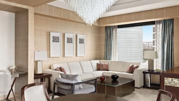 A modern living room with a white sofa, a glass coffee table, and a chandelier. There are framed pictures on the wall and a view of the city through large windows.