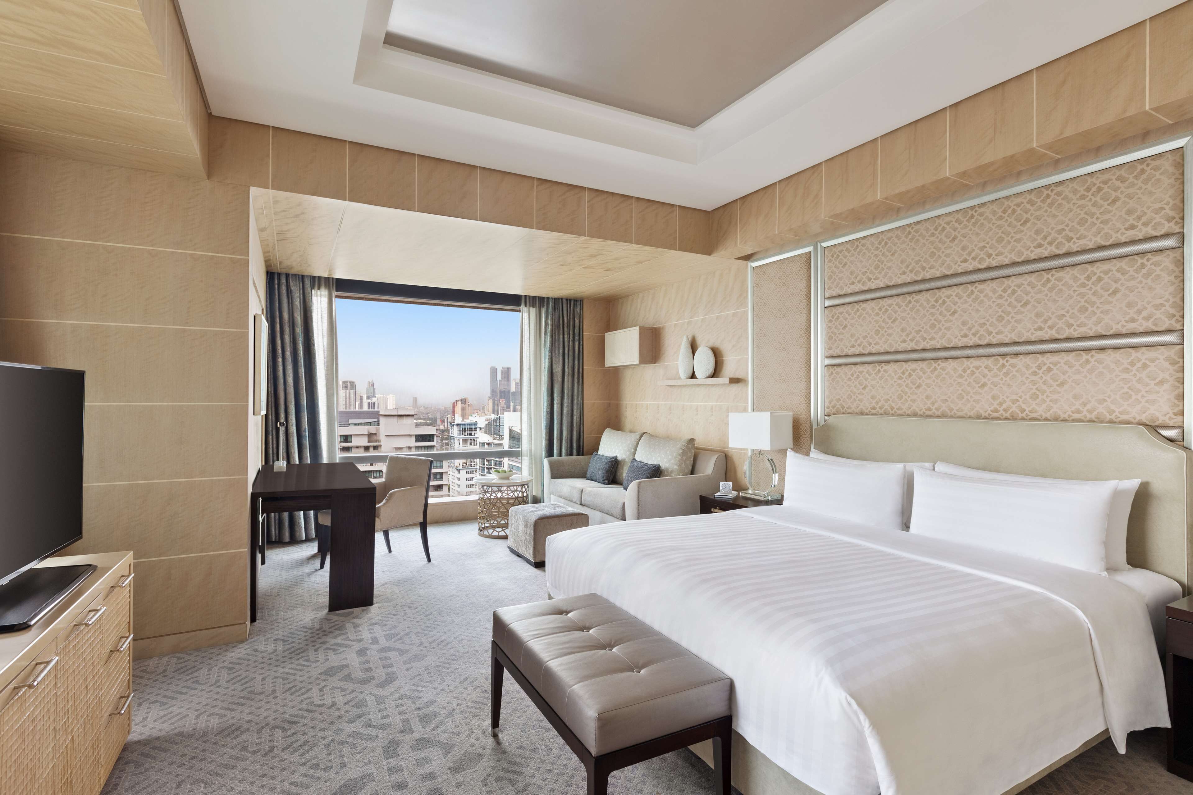 A modern hotel room with a large bed, a desk, a sofa, and a view of the city.