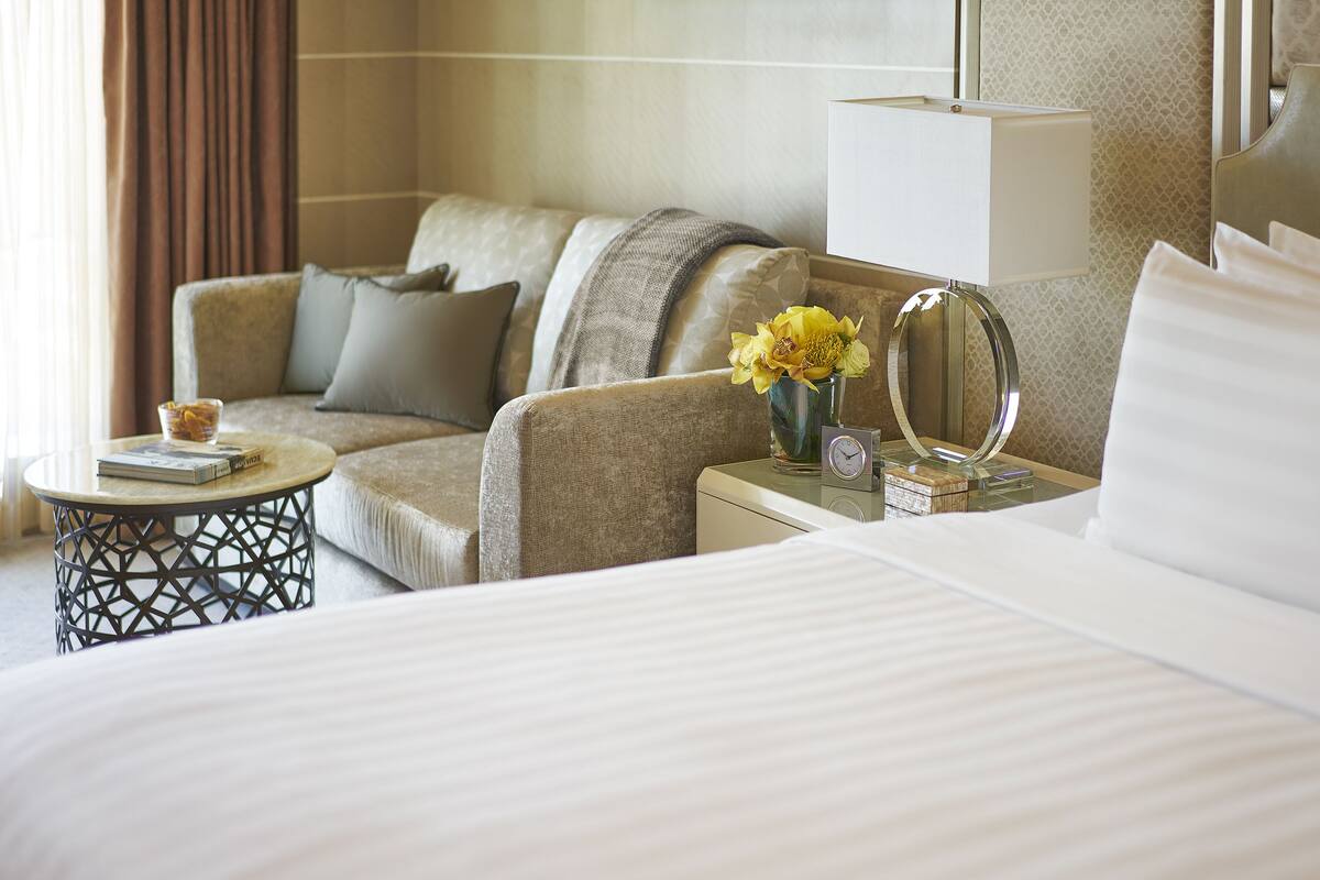 Une chambre d’hôtel avec un lit, un canapé, une petite table de chevet avec une lampe, un vase de fleurs et une horloge.