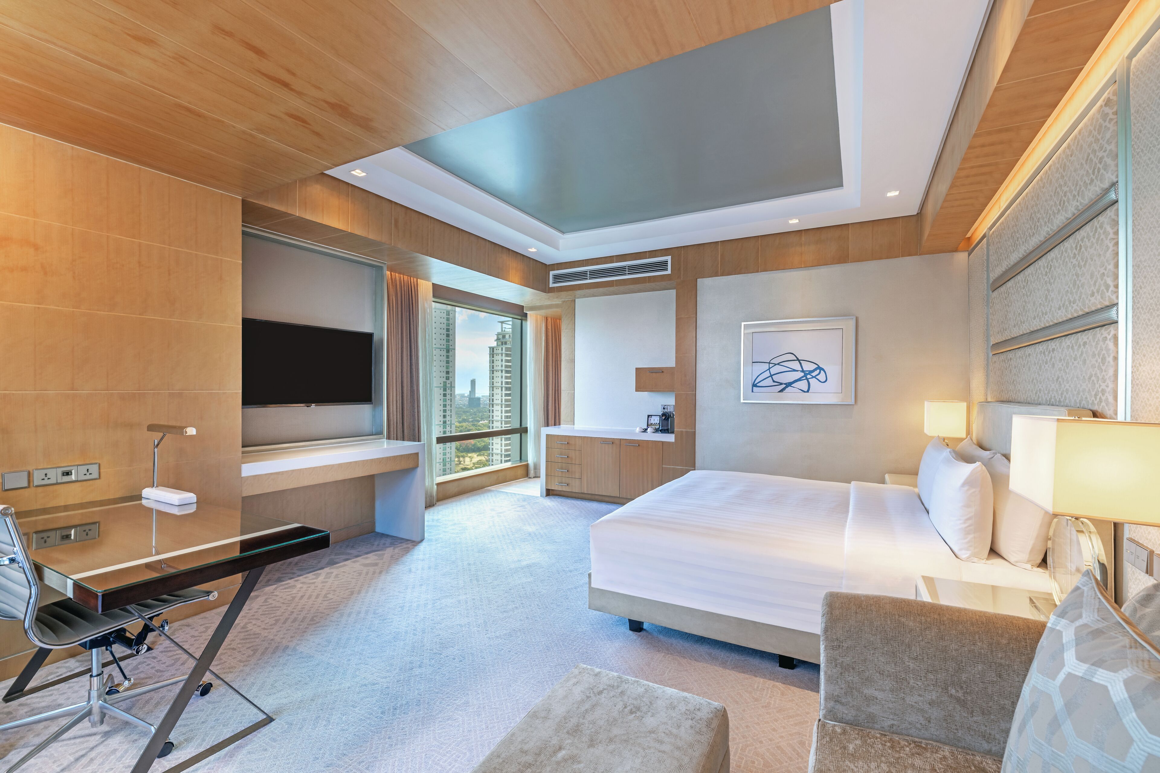 A modern hotel room with a bed, desk, chair, and a large window offering a city view.