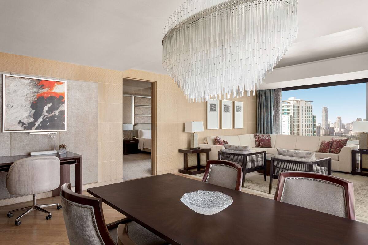 Une chambre d’hôtel moderne dotée d’un coin repas, d’un ensemble de canapés, d’un lustre et offrant une vue sur la ville.