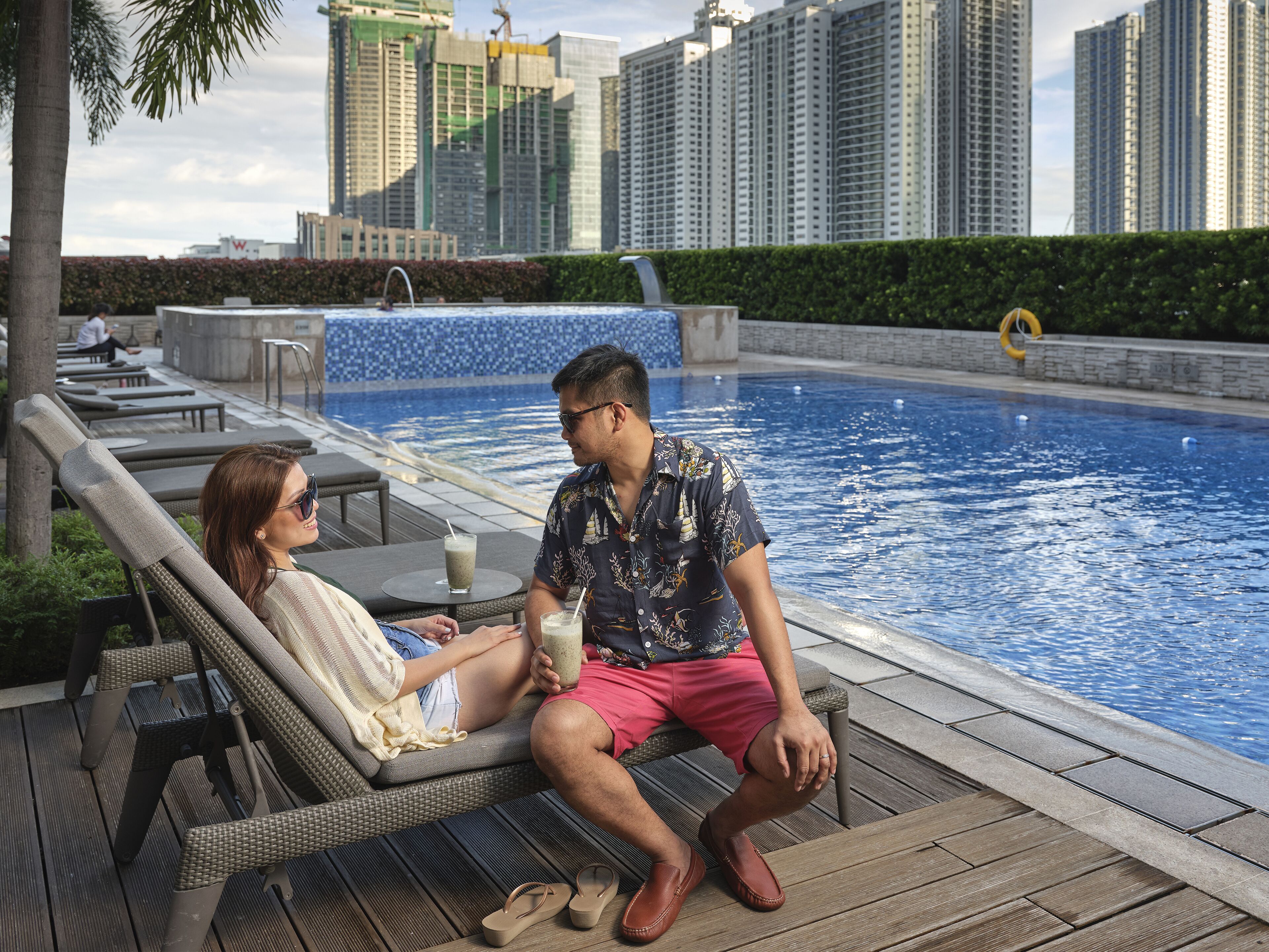 Un área junto a la piscina con sillones de descanso, una piscina y edificios altos al fondo.