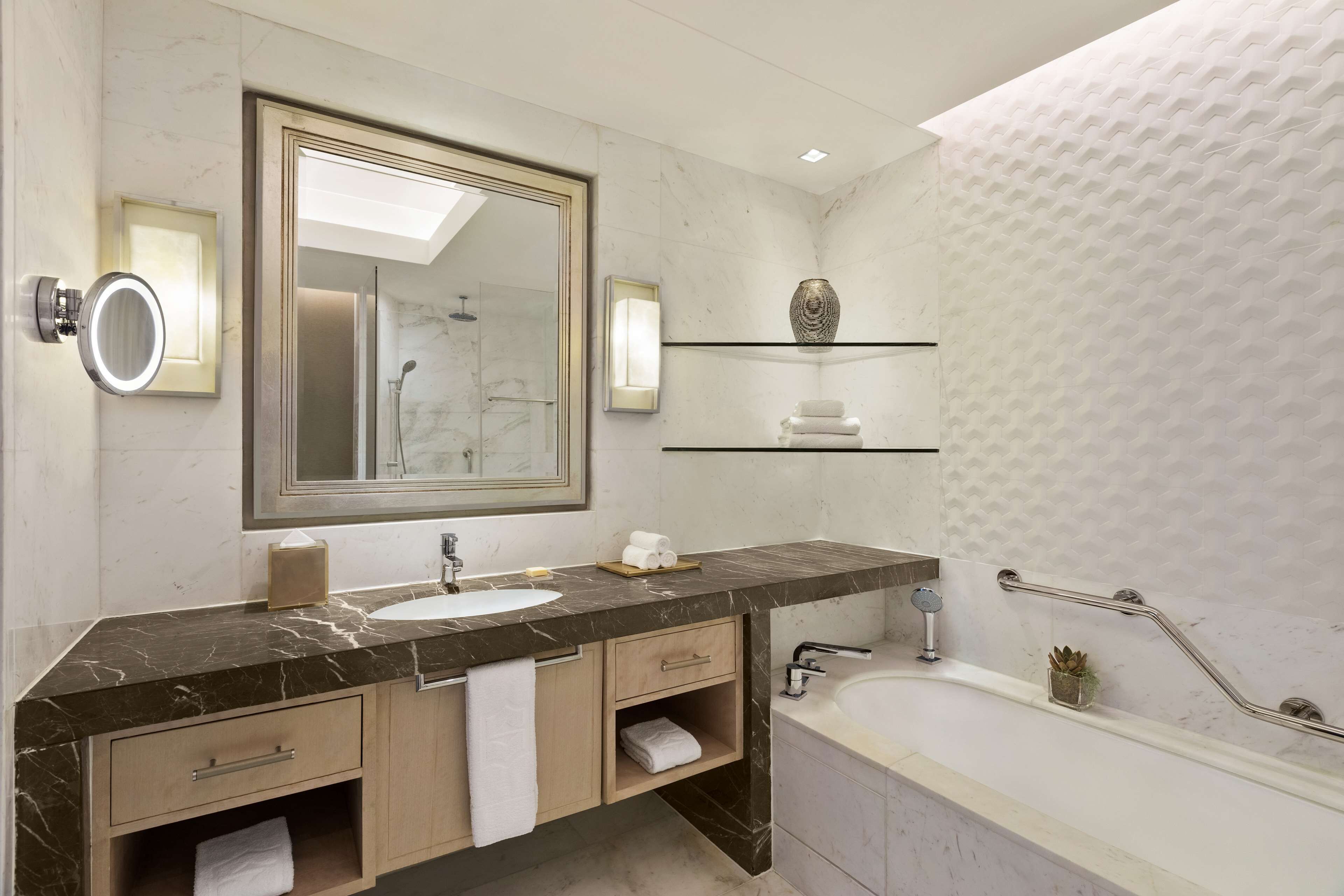 A modern bathroom with a marble vanity, a large mirror, a built-in sink, a bathtub, and a shelf with toiletries.