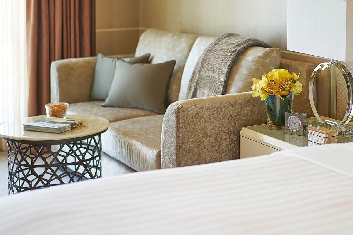 A hotel room with a bed, a sofa, a small table with a clock, and a vase with flowers.
