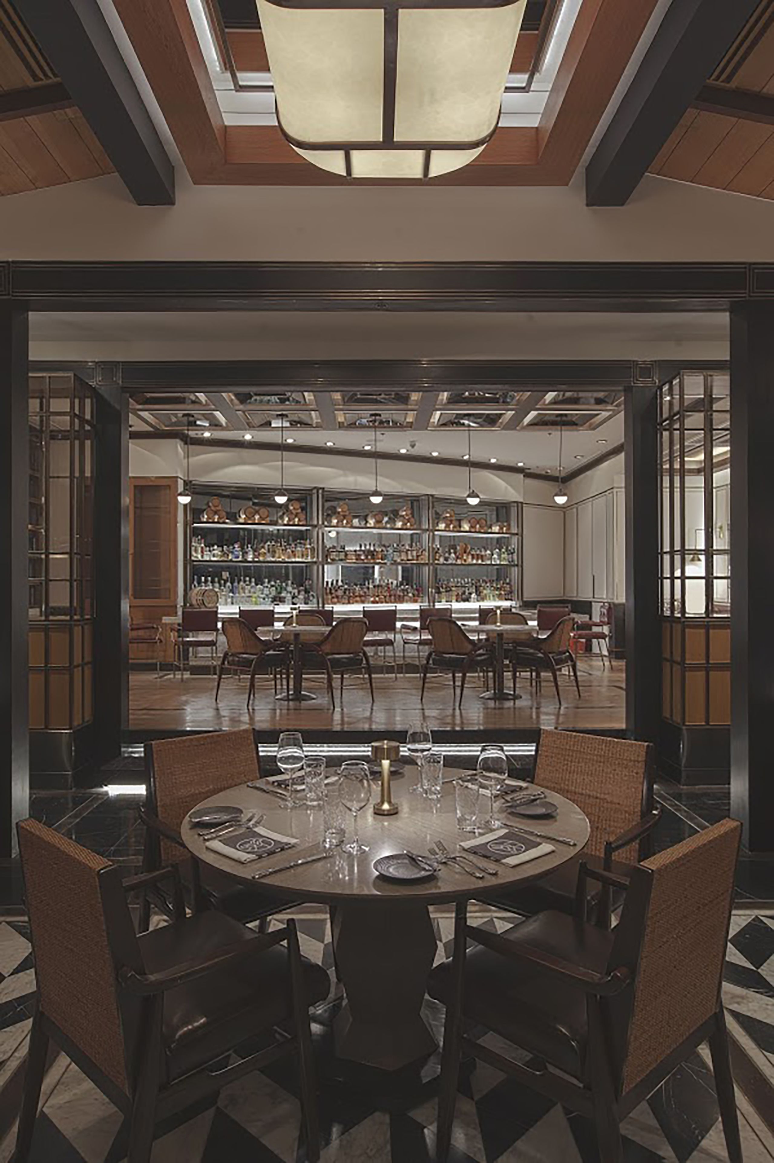 A dining area with a round table set for four, surrounded by wicker chairs, with a bar and shelves of bottles in the background.