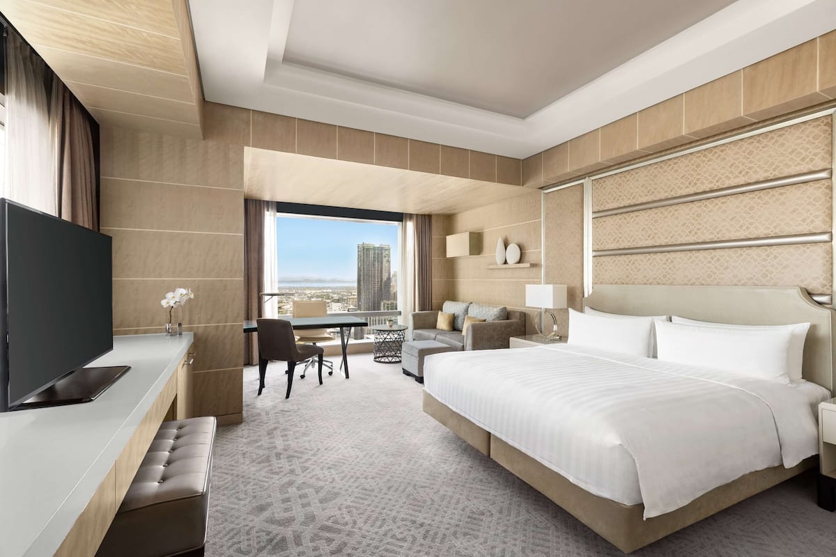 Une chambre d’hôtel moderne dotée d’un grand lit, d’un bureau avec un téléviseur à écran plat, d’un coin salon avec un canapé et des fauteuils, et offrant une vue sur le paysage urbain.