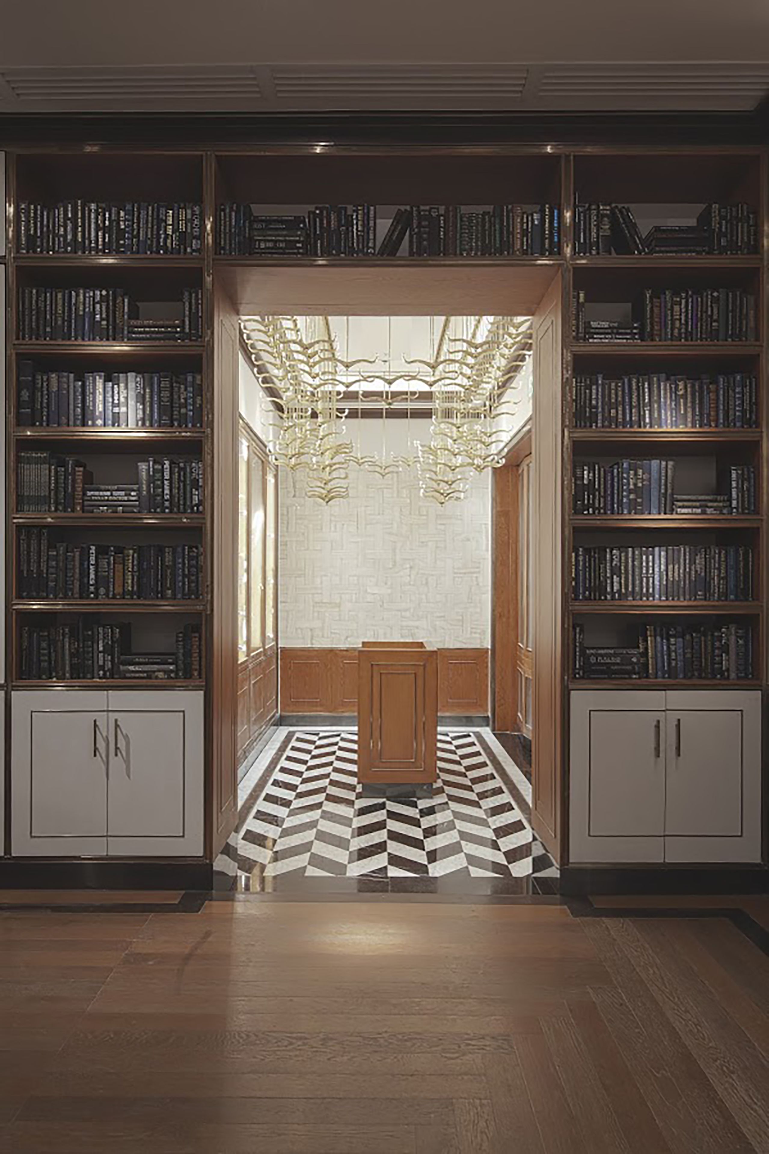 A library with bookshelves on both sides, a central reading area with a patterned floor, and a chandelier.