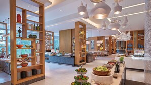 A modern hotel lobby with wooden shelving displaying various kitchenware, a central bar area with a chef preparing food, and a buffet with fresh fruits and vegetables. - Shangri-La The Fort, Manila (Taguig)