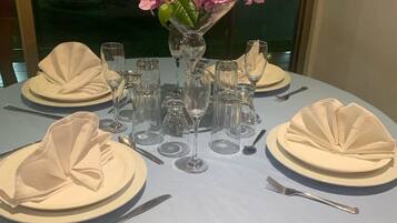 Een ronde eettafel gedekt voor vier personen, met witte tafelkleden, glazen en roze bloemen in een vaas.
