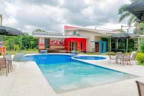 Una piscina con cascada, rodeada de mobiliario exterior y un edificio de fachada roja y azul.