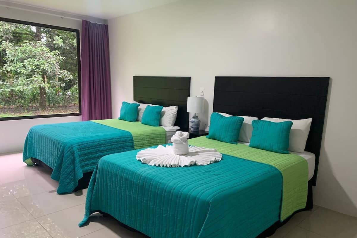Habitación de hotel con dos camas, cada una con ropa de cama color turquesa y almohadas blancas. Se aprecia una ventana con vista a árboles y una cortina morada.
