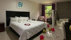 A hotel room with a large bed, a small desk, a chair, a bathtub, and a view of greenery outside.