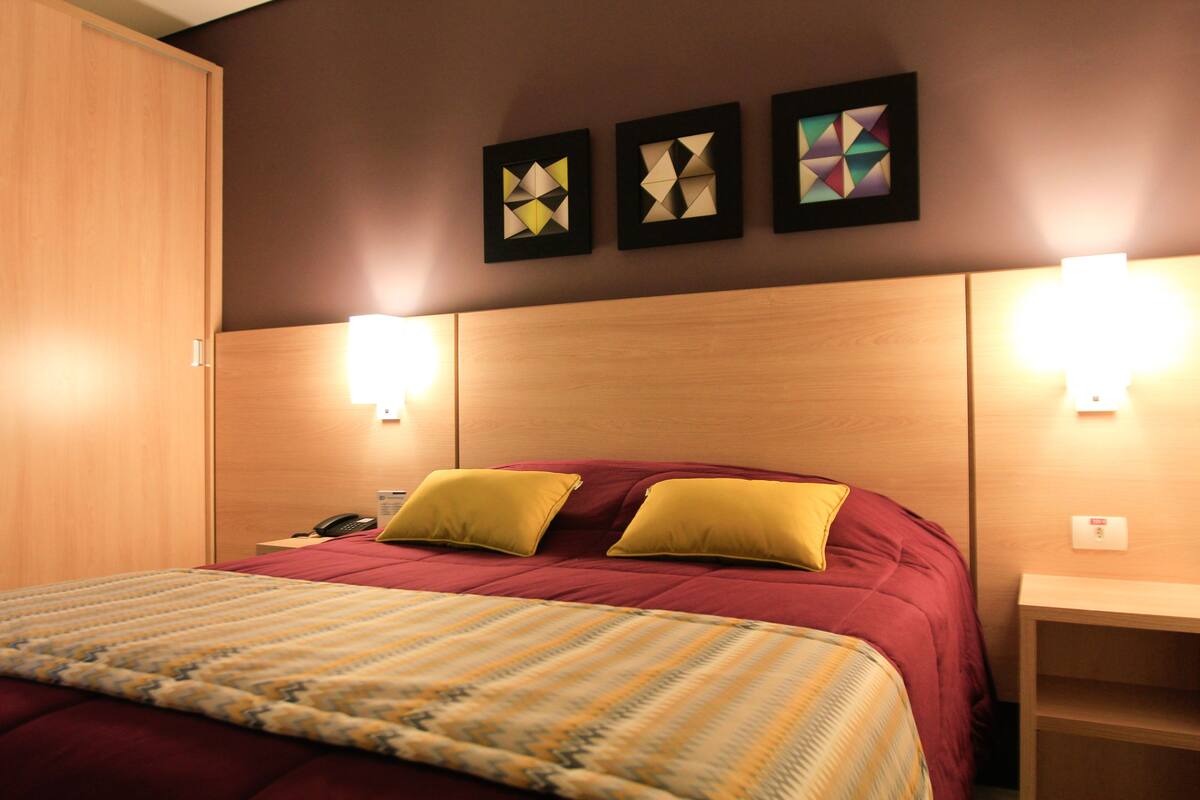A hotel room with a large bed, two yellow pillows, a wooden headboard, and three framed geometric wall art pieces.