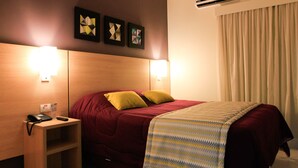 Habitación de hotel con cabecera de madera, cubrecama rojo, dos almohadas amarillas, manta decorativa estampada y dos lámparas fijadas a la pared.