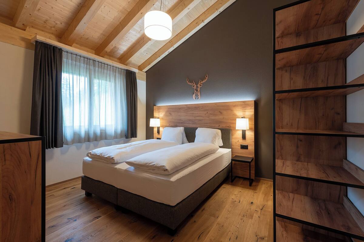 Uma cama de casal em quarto com vigas de madeira, decoração de cabeça de veado e mesa de cabeceira com abajur.
