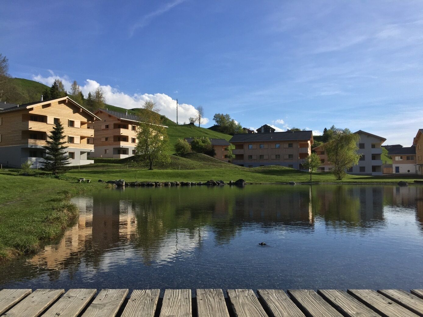 Vista sobre un llac amb edificis de fusta, un moll i un cos d'aigua tranquil·la que reflecteix les construccions.