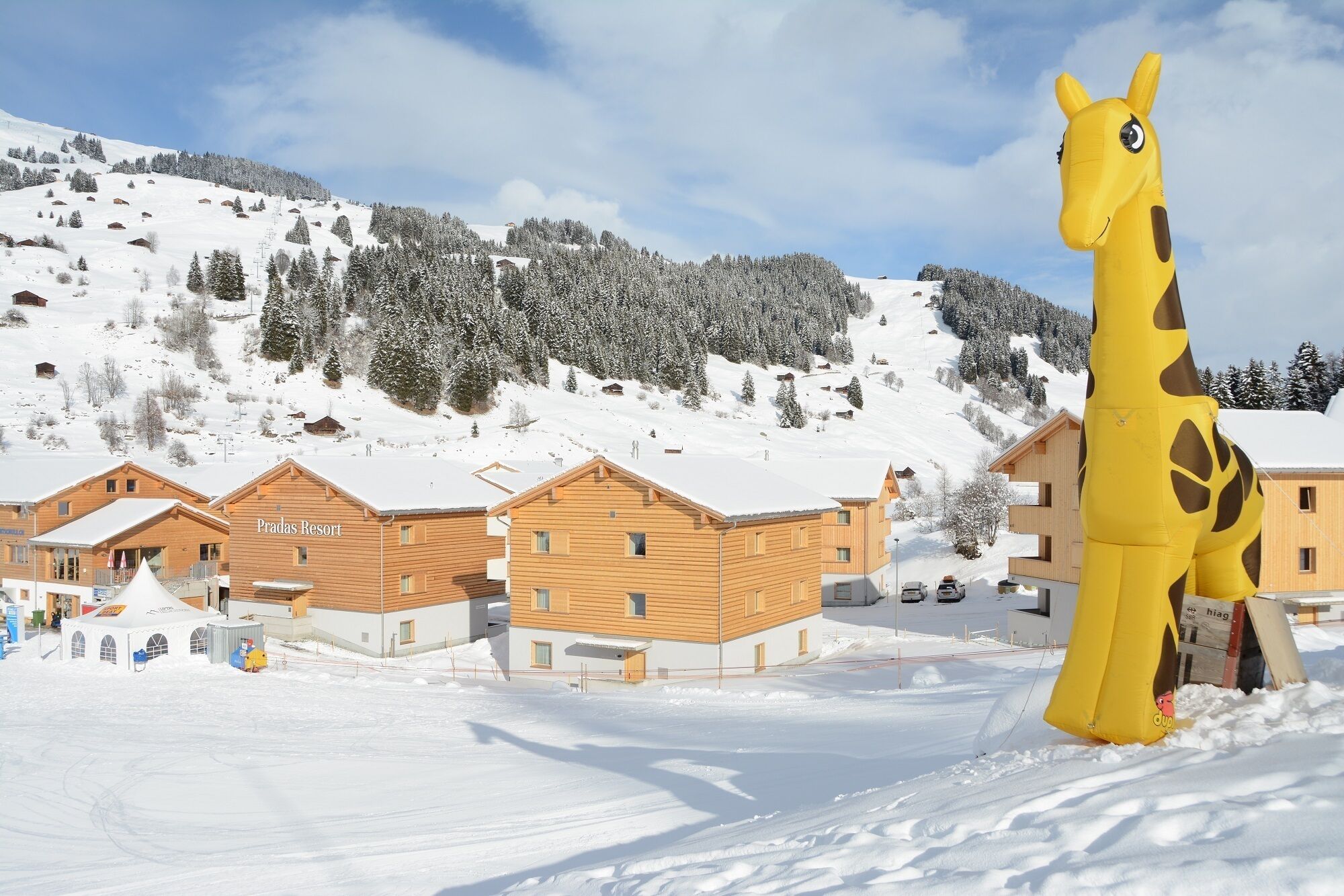Une station de ski enneigée, avec des chalets en bois et une grande décoration gonflable en forme de girafe.