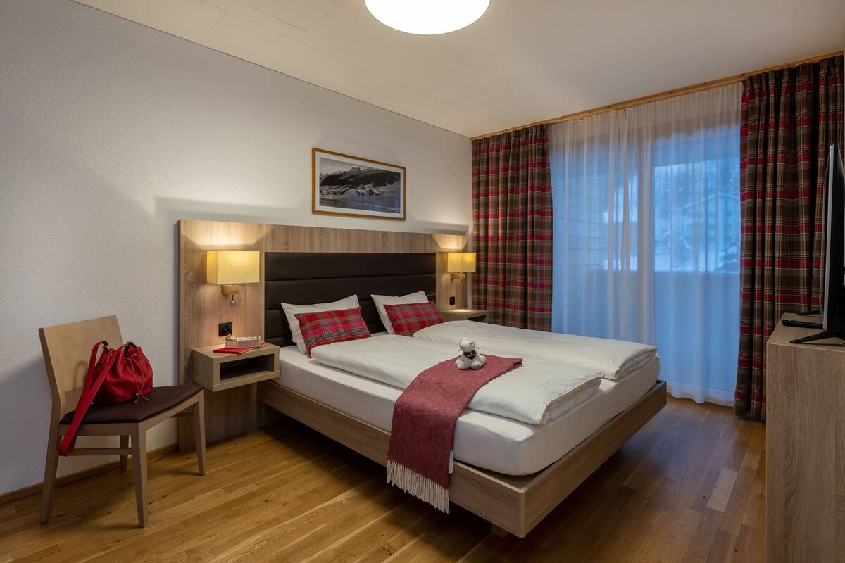 Quarto de hotel moderno com uma cama grande, cabeceira de madeira, duas mesas de cabeceira com abajures, uma cadeira vermelha, uma bolsa vermelha e um quadro na parede.
