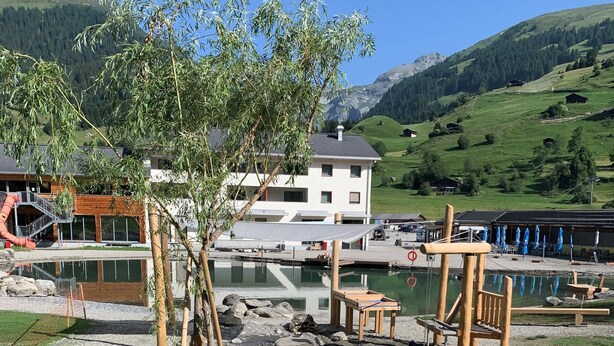 A riverside area with a wooden boat, a building with a white facade, and a mountain in the background. - Pradas Resort Brigels (Brigels)