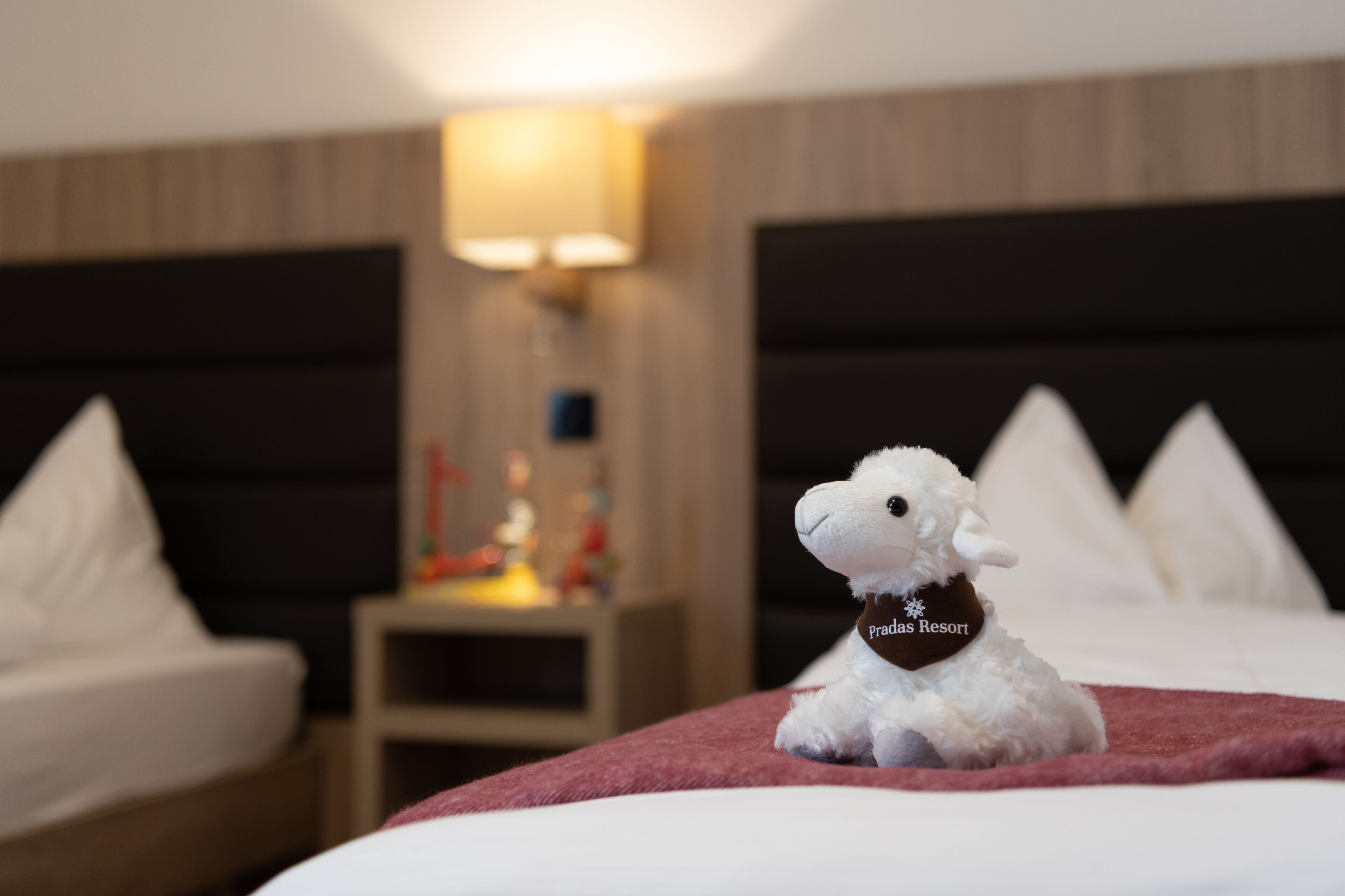 Ein flauschiges weißes Schafspielzeug auf einem Bett mit einer roten Decke, einem Nachttisch mit einer Lampe und einem Beistelltisch mit einem kleinen dekorativen Gegenstand.
