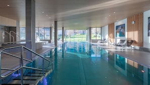 A modern indoor swimming pool with clear glass walls, a view of a green landscape, and lounge chairs along the poolside. - Pradas Resort Brigels (Brigels)