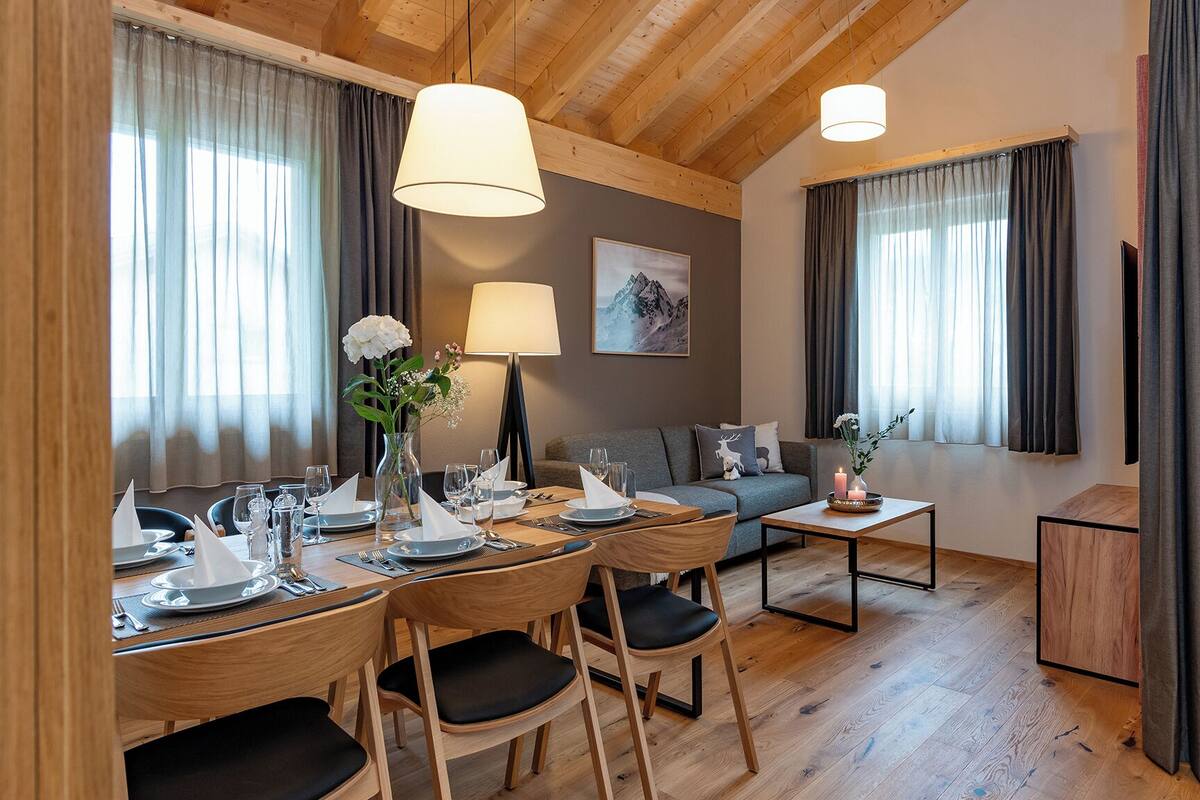 Uma área de refeições com uma mesa de madeira posta para quatro pessoas, um sofá e uma pequena mesa lateral com velas.