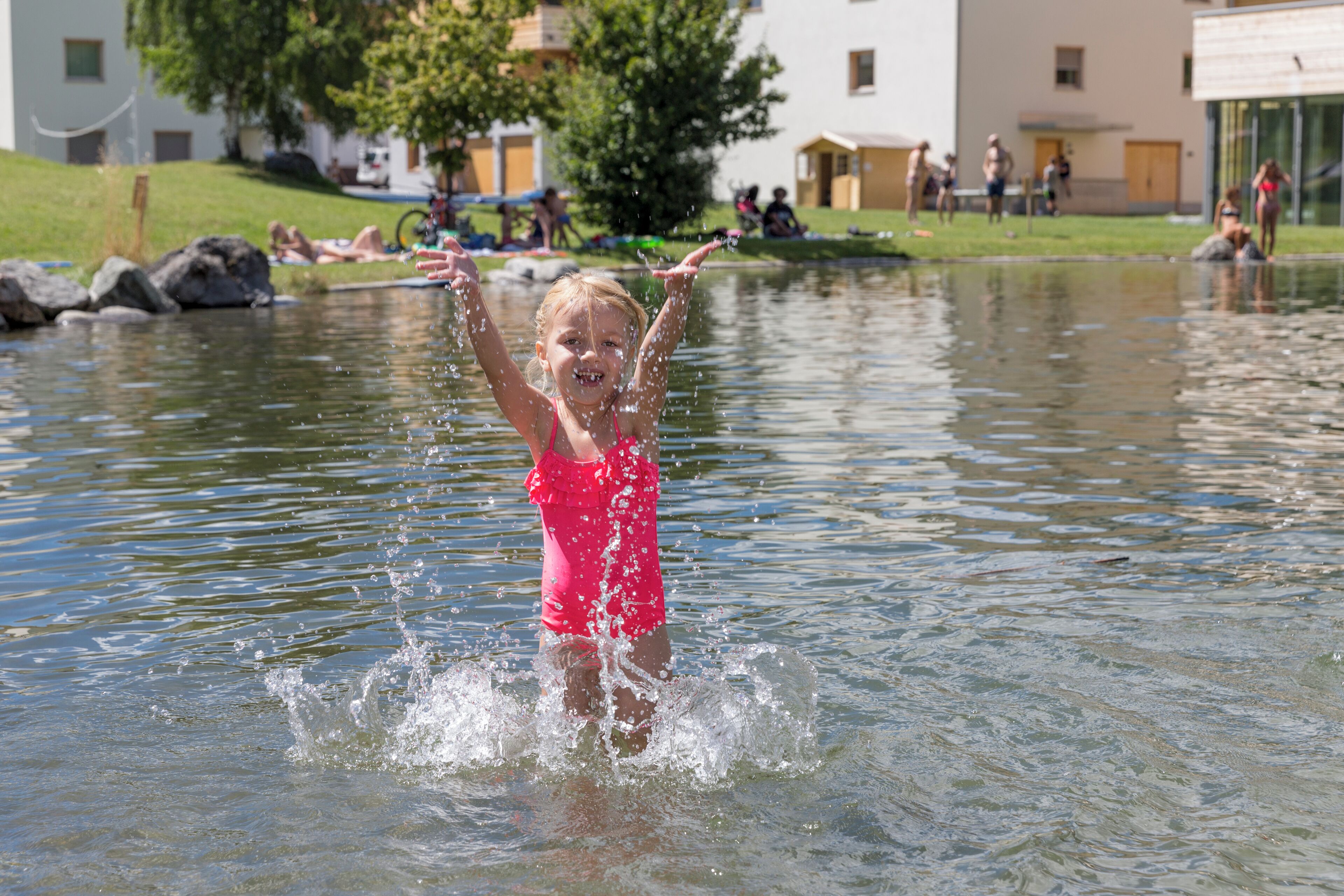 Een kind speelt in het water met zicht op gebouwen en een grasveld op de achtergrond.