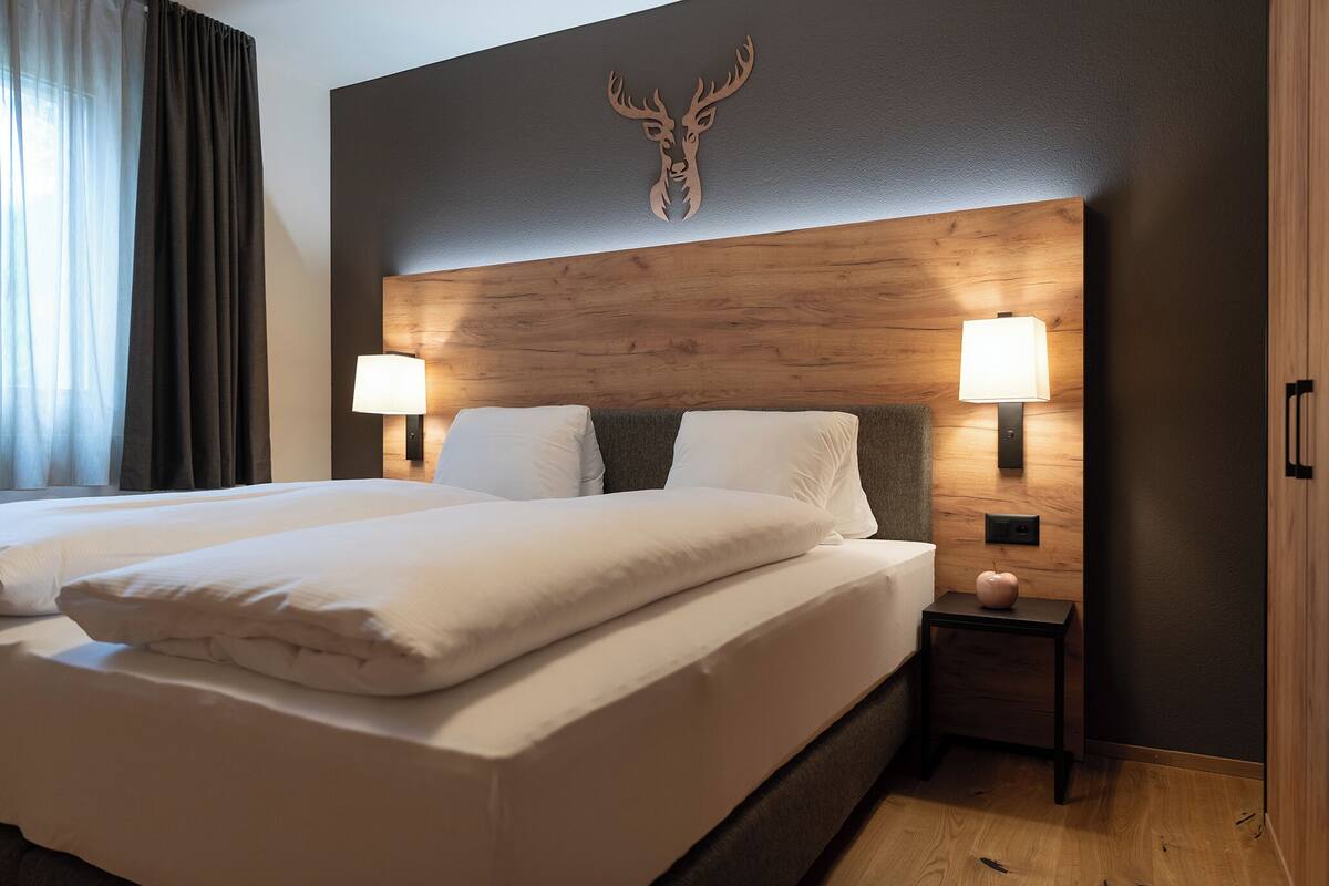 Cama de casal com roupa de cama branca, duas luminárias de cabeceira, mesa de cabeceira de madeira pequena com uma luminária e decoração de cabeça de veado na parede.