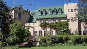 Un grand bâtiment imposant, qui ressemble à un château, avec de nombreuses tours et des drapeaux, entouré de jardins bien entretenus et sous un ciel dégagé.