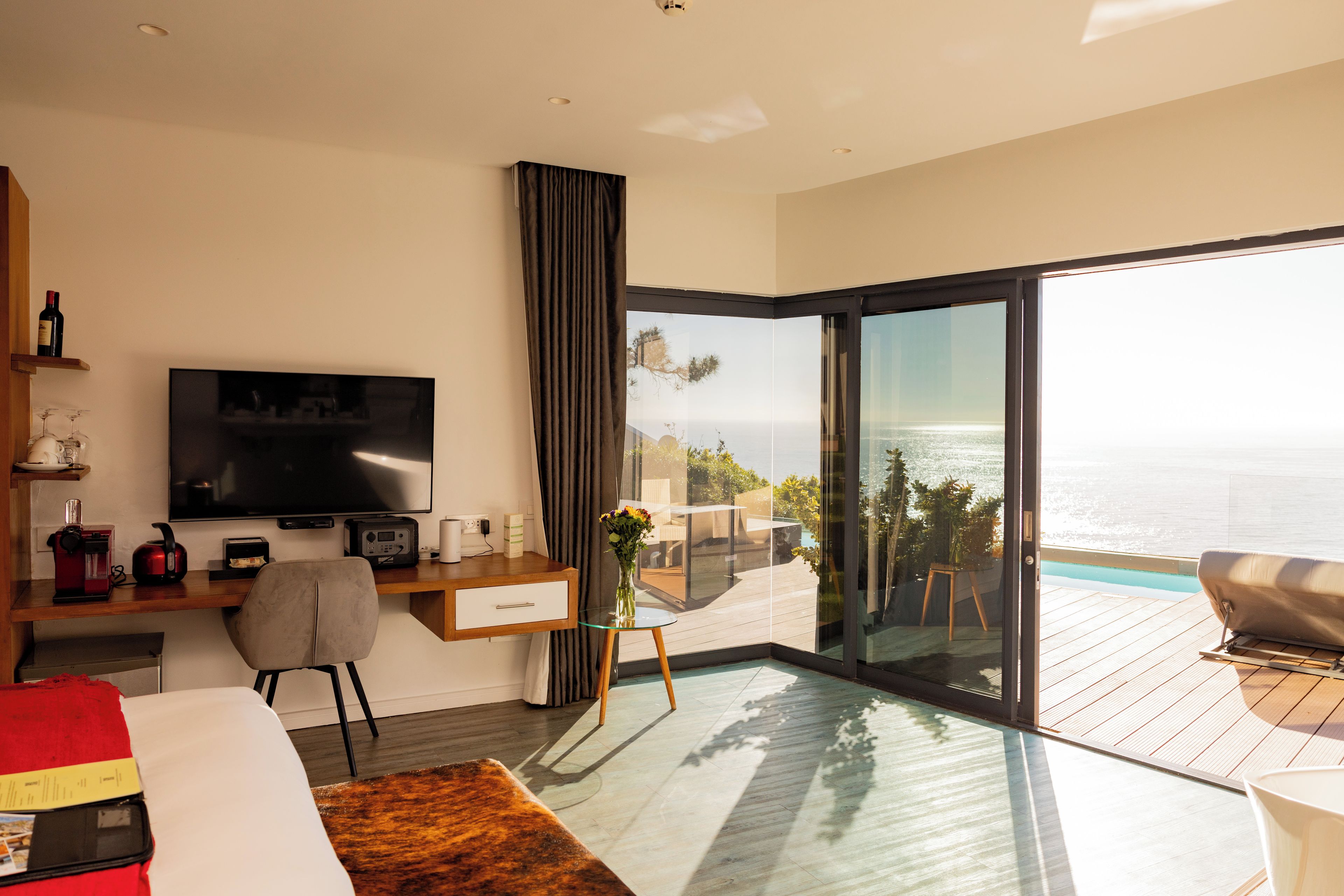 Ein modernes Hotelzimmer mit großem Fernseher, einem Schreibtisch inklusive Computer, einem bequemen Sessel, einem kleinen Tisch mit Blumenvase und einem Balkon mit Meerblick.