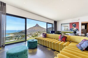 A modern living room with a yellow sofa, teal ottoman, and a large window offering a view of a mountain and ocean. - Sea Star Rocks (Cape Town)