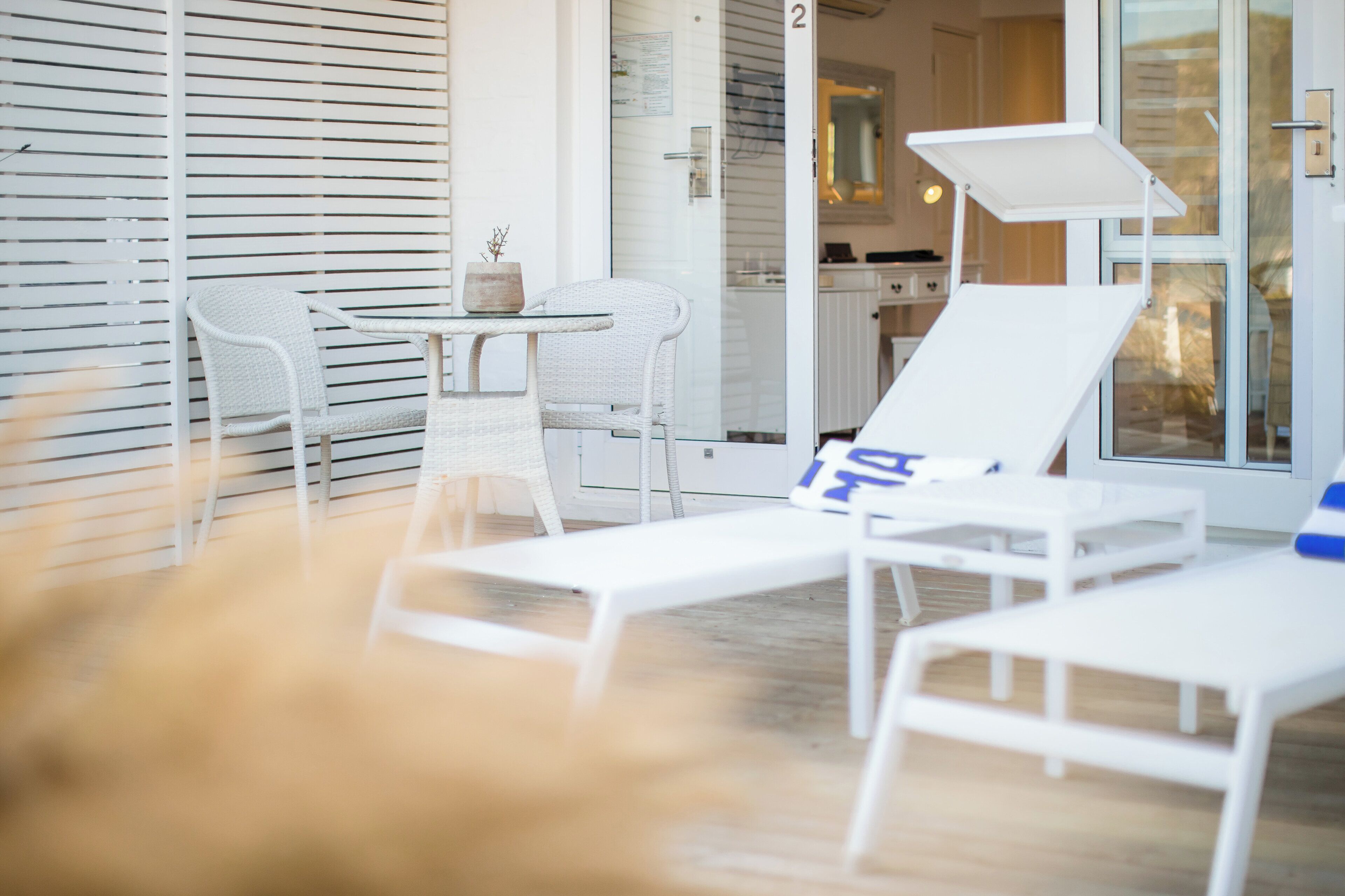 A modern outdoor lounge area with white furniture, including two lounge chairs and a table, near a sliding glass door leading to a kitchen area.