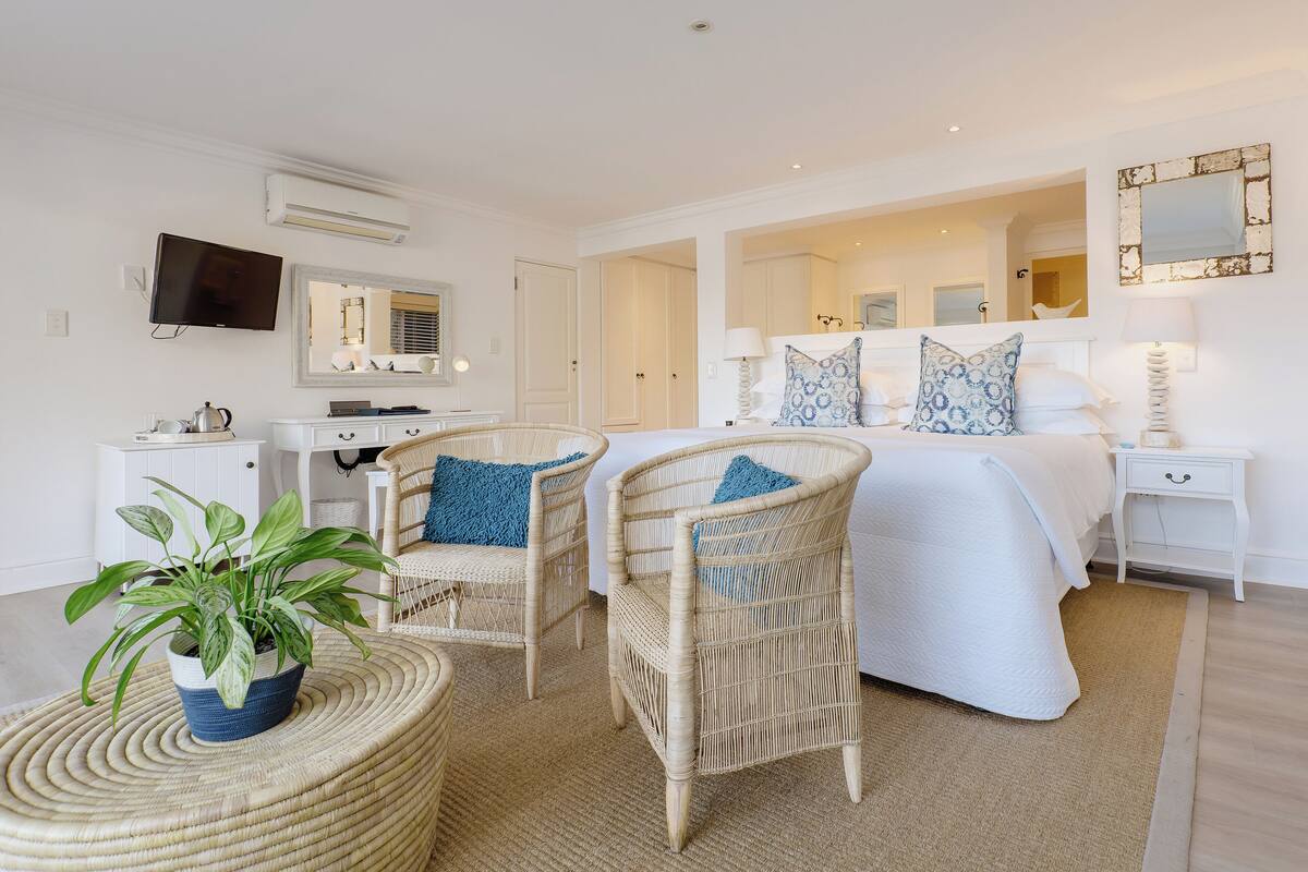A hotel room with a large bed, two wicker chairs, a small table with a plant, a TV, and a mirror.