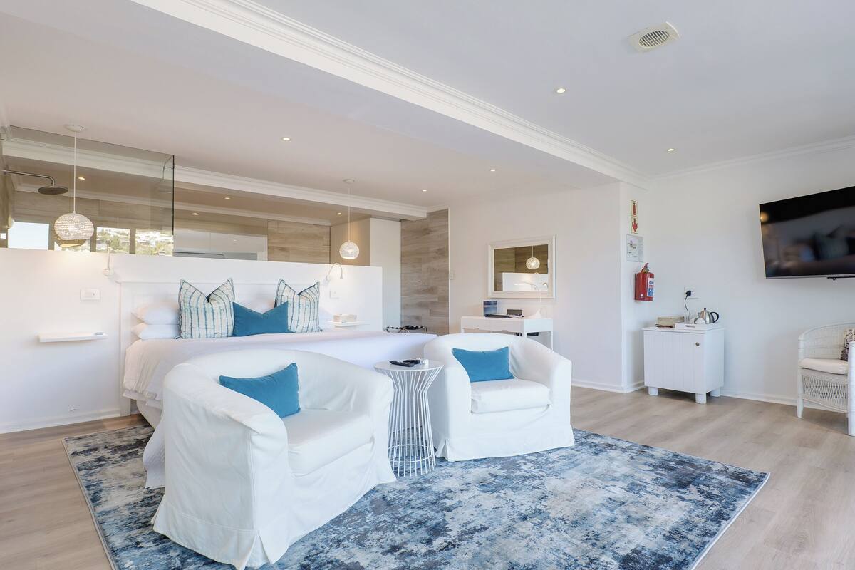 A modern bedroom with a large bed, two white armchairs, a small round table, and a large mirror.