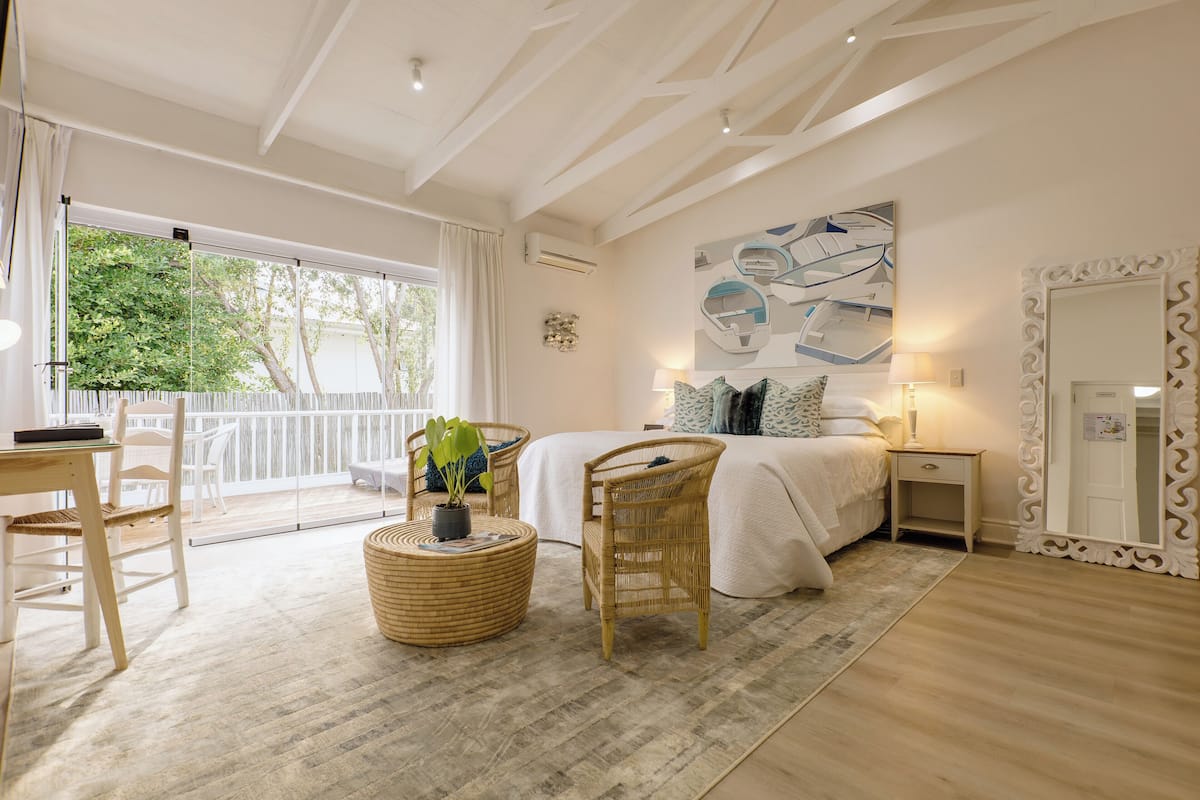 A bedroom with a large bed, a bedside table, a mirror, and a view of the outdoors.