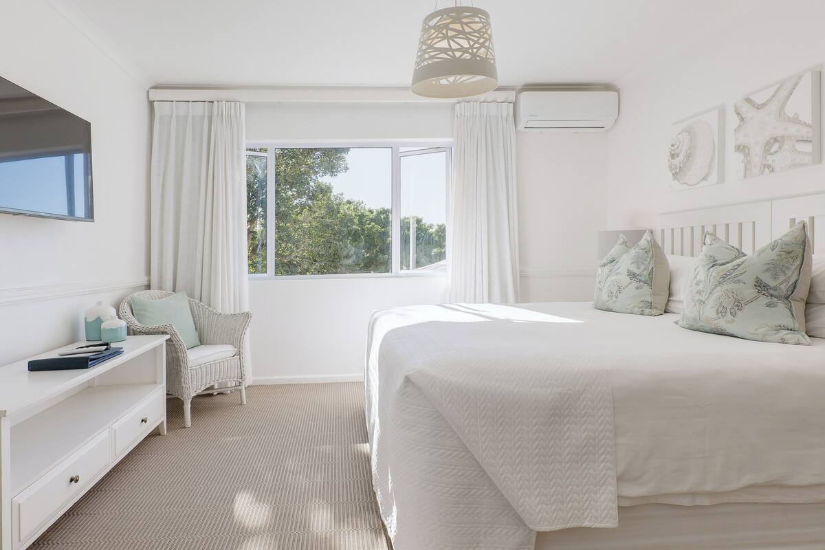 A bedroom with a large bed, a bedside table, a chair, a window with curtains, and wall art.