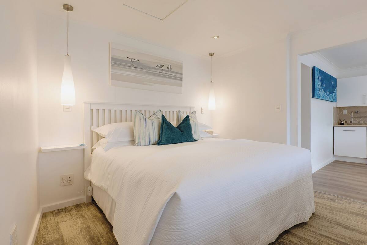 A bedroom with a large bed, white walls, a wooden floor, and a kitchen area visible through an open doorway.