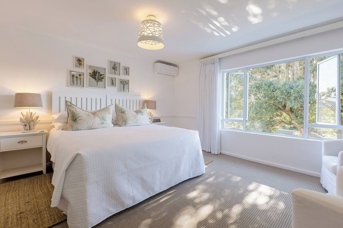 A bedroom with a large bed, bedside tables, a lamp, framed botanical prints on the wall, and a window with a view of greenery.