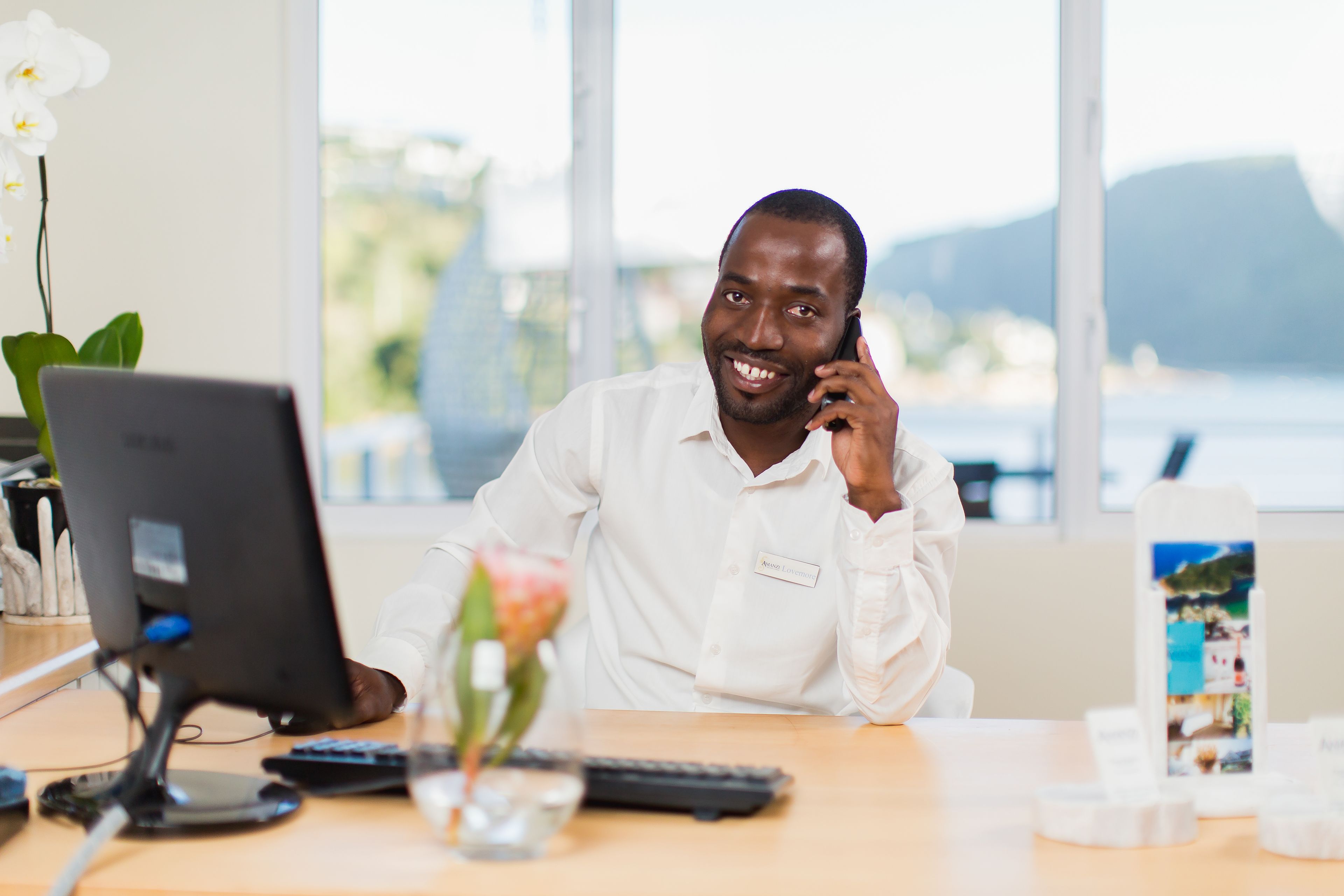 A man at a desk with a computer, a vase with flowers, and a view of the outdoors.