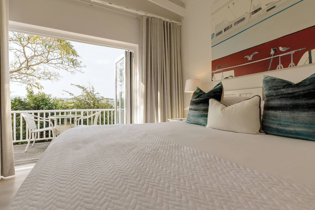 A spacious bedroom with a large bed, a balcony with outdoor furniture, and a view of greenery.
