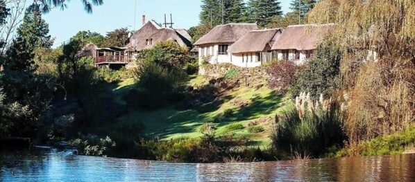 A riverside property with thatched-roof cottages, a grassy embankment, and a calm water body. - Hawklee Country House (Fort Nottingham)