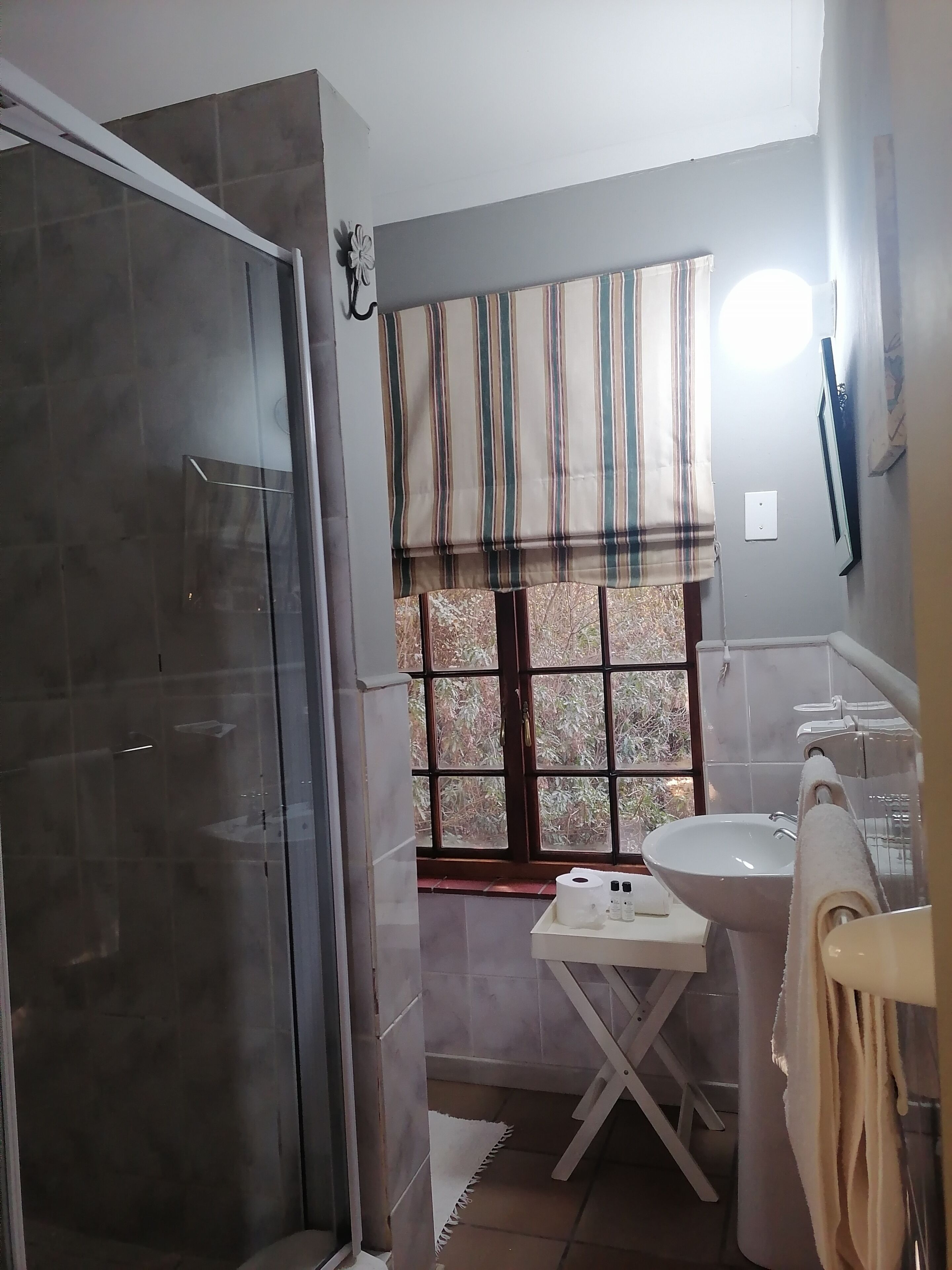 A bathroom with a shower, a window with striped curtains, a sink, and a towel rack.