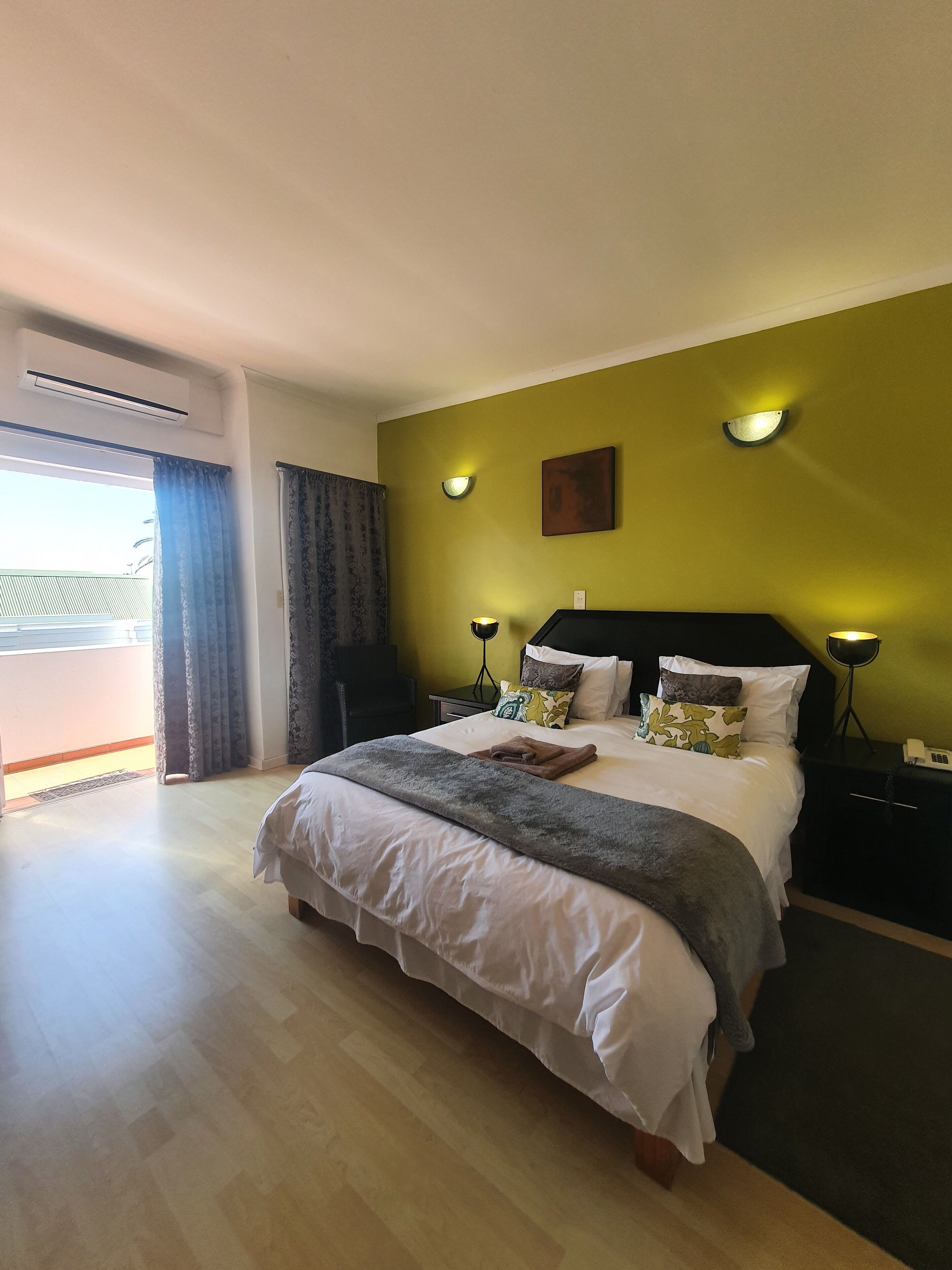 Una habitación de hotel con cama, mesitas de noche, lámpara de pared y ventana con cortinas.